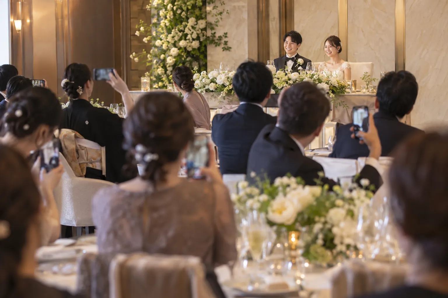 ホテルの披露宴で、円卓のゲストがスマートフォンで新郎新婦を撮影している様子