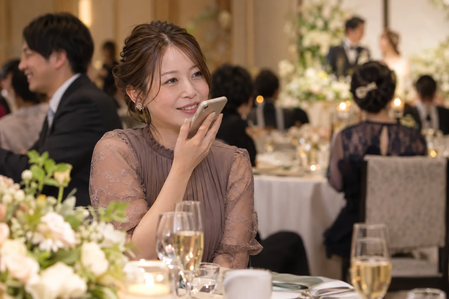 披露宴の円卓で、女性ゲストがスマートフォンに音声メッセージを吹き込んでいる様子