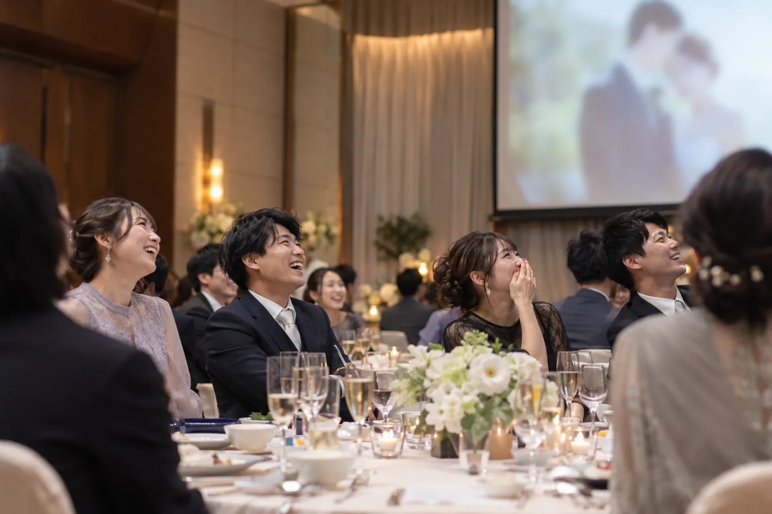 披露宴の円卓で、ゲストが笑いながら会場のスクリーンを見上げている様子